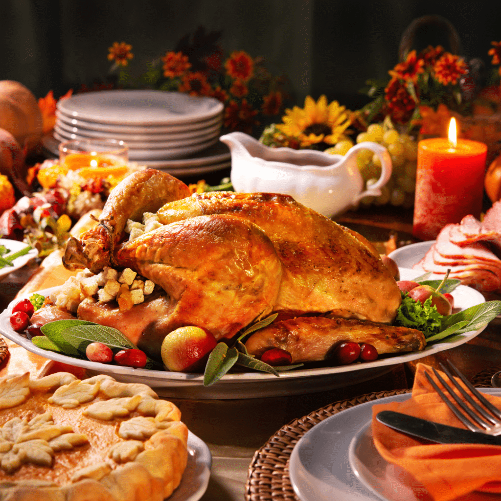 Festive dinner table setup for Thanksgiving. The centerpiece is a roasted turkey garnished with herbs, apples, and cranberries. Surrounding the turkey are various dishes, including a pie with leaf-shaped crust decorations, a stack of plates, a gravy boat, a lit candle, and a platter of sliced ham. The table is decorated with autumn-themed elements such as flowers and pumpkins, creating a warm and inviting atmosphere.